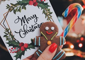 Close-up of a hand holding a gingerbread man, candy cane, and Merry Christmas card in front of a decorated tree.